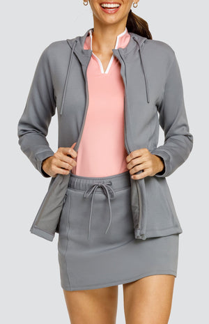 Model wearing a gray hooded jacket with a cinched waist and drawstring hood. Paired with a matching skort with drawstring waistband, and a light pink top with white accents.