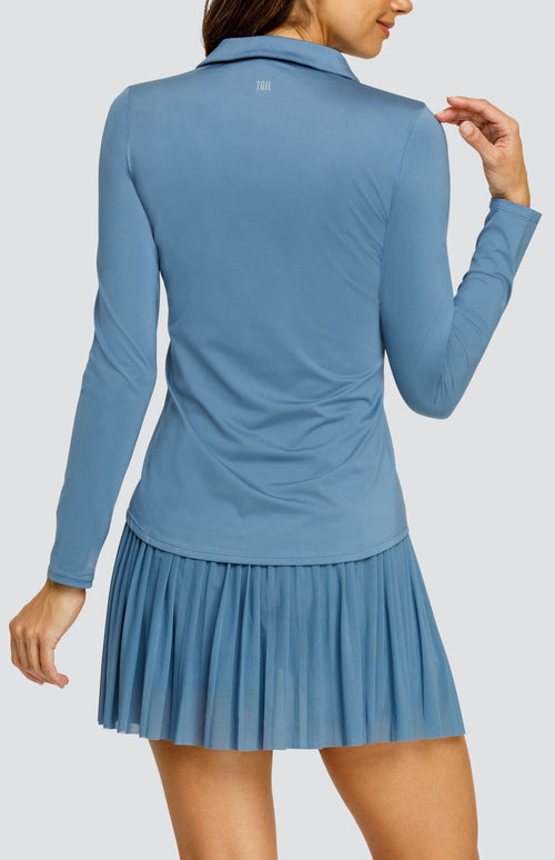 Model wearing a steel blue long sleeve top with an open v neck foldover collar with white accents and side vents. Paired with a matching mesh pleated skort with solid undershorts.