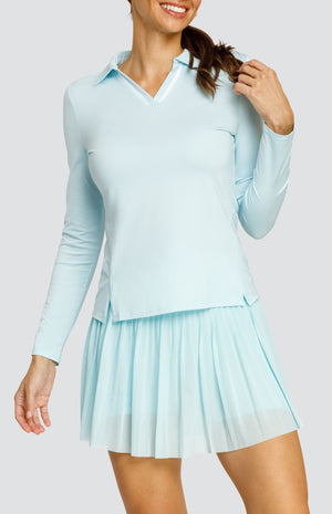 Model wearing a light blue long sleeve top with an open v neck foldover collar with white accents and side vents. Paired with a matching mesh pleated skort with solid undershorts.