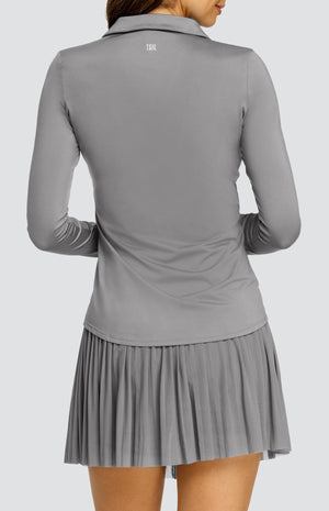 Model wearing a gray long sleeve top with an open v neck foldover collar with white accents and side vents. Paired with a matching mesh pleated skort with solid undershorts.