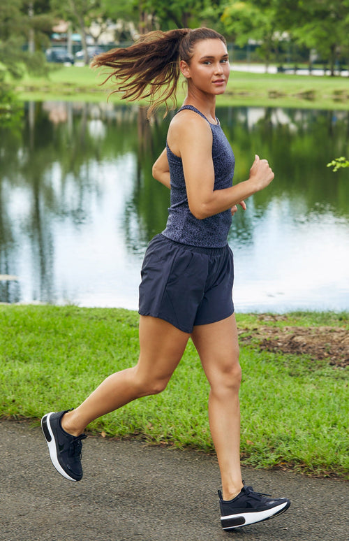 Woman running next to a lake wearing a high neck tank top in a mottled gray leopard print and black shorts with an elastic waistband and pockets.