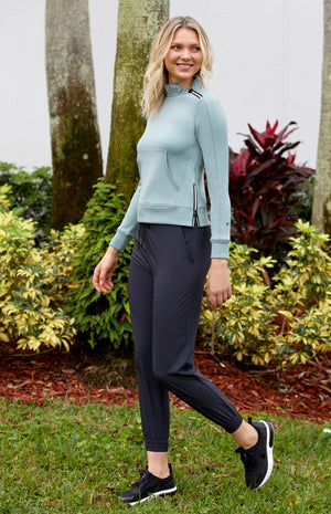 Woman standing in a park wearing a sage colored pullover with a ruffled collar, zippers, and a kangaroo pocket, and wearing black joggers with elastic drawstring waistband and elastic cuffs.