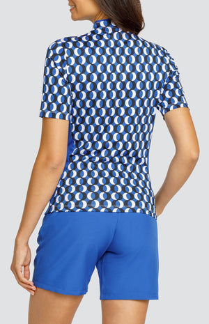 Model wearing a short sleeve golf top with a circular blue, white, and black pattern, and blue shorts.