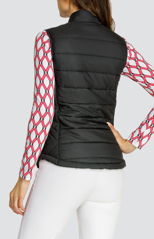 Model wearing a black puffer vest over a long sleeve quarter-zip golf top with a pink and white lattice pattern and a pull on pink golf skort with a flounce insert.