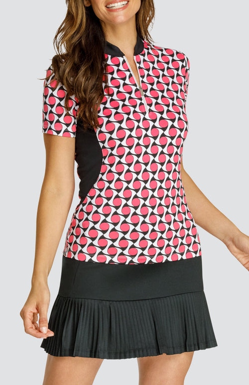 Model wearing a short sleeve quarter-zip golf top with a pink, white and black geometric pattern and black inserts, with a black pleated golf skort.