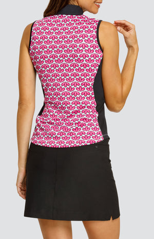 Model wearing a sleeveless quarter-zip golf top in a pink geometric floral print on a white background with black side inserts, paired with a solid black pull-on golf skort.