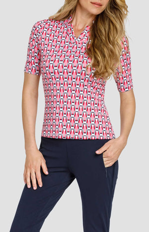 Front view of a woman wearing a mid-length sleeve golf top with a star collar neckline. The print is a geometric pattern of 6-sided gemstones in a light pink color on a white background. She is also wearing dark navy blue pants with zipper pockets.