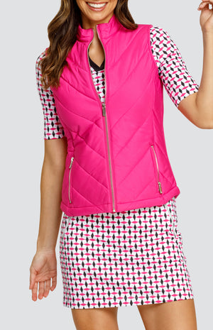 Model wearing an open neck elbow sleeve golf top in a pink and black geometric alternating diamond print on a white background with black side inserts, paired with a pull-on skort in a matching print and a solid pink zip-up puffer vest.