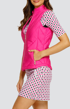 Model wearing a solid pink zip-up puffer vest, paired with an open neck elbow sleeve golf top in a pink and black geometric alternating diamond print on a white background with black side inserts and a pull-on skort in a matching print.
