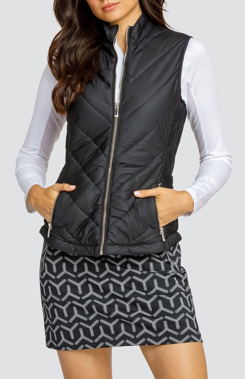 Model wearing a white raglan long sleeve golf top and a black and white geometric print skort, and a black sleeveless puffer vest.