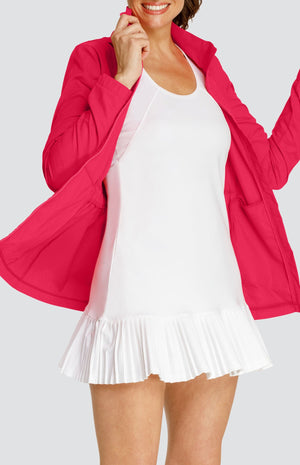 Model wearing a pinkish red water resistant jacket with a drawstring waist and attached hood, over a white tennis dress with pleated hem.