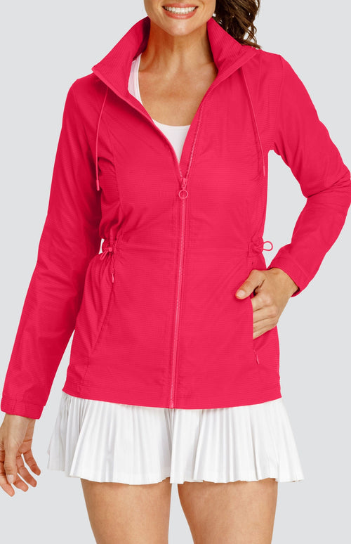 Model wearing a pinkish red water resistant jacket with a drawstring waist and attached hood, over a white tennis dress with pleated hem.