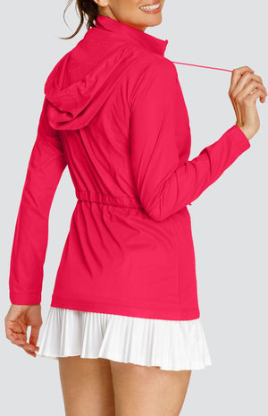 Model wearing a pinkish red water resistant jacket with a drawstring waist and attached hood, over a white tennis dress with pleated hem.