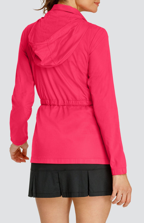 Model wearing a pinkish red water resistant jacket with a drawstring waist and attached hood, over a black pleated skort.